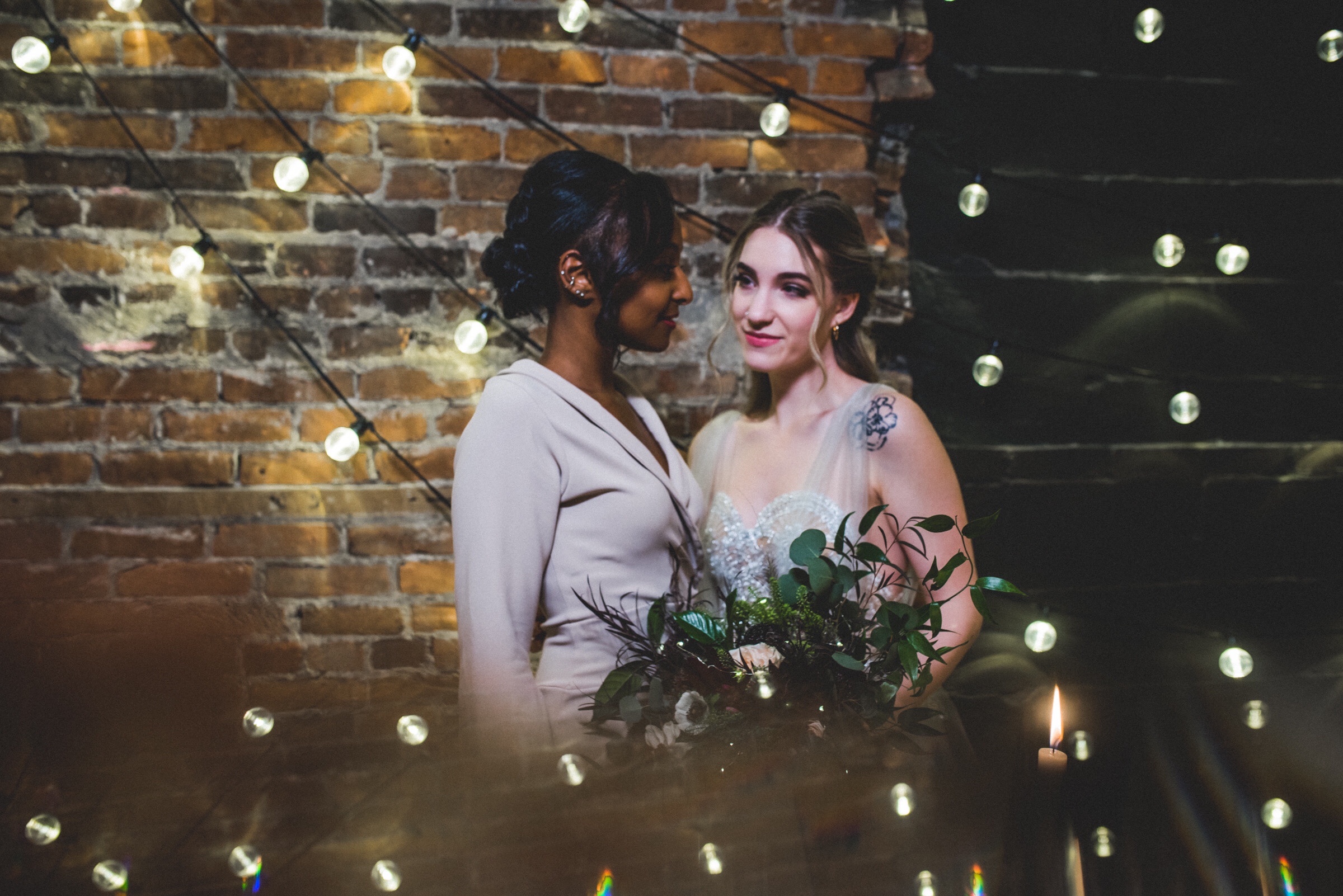 Same sex bridal couple surrounded by lights at minnesota wedding venue The loft at studio J. Portrait captured by minnesota wedding photographer Madelin Zaycheck of the Hawk and Sparrow using a prism for reflection of the lights